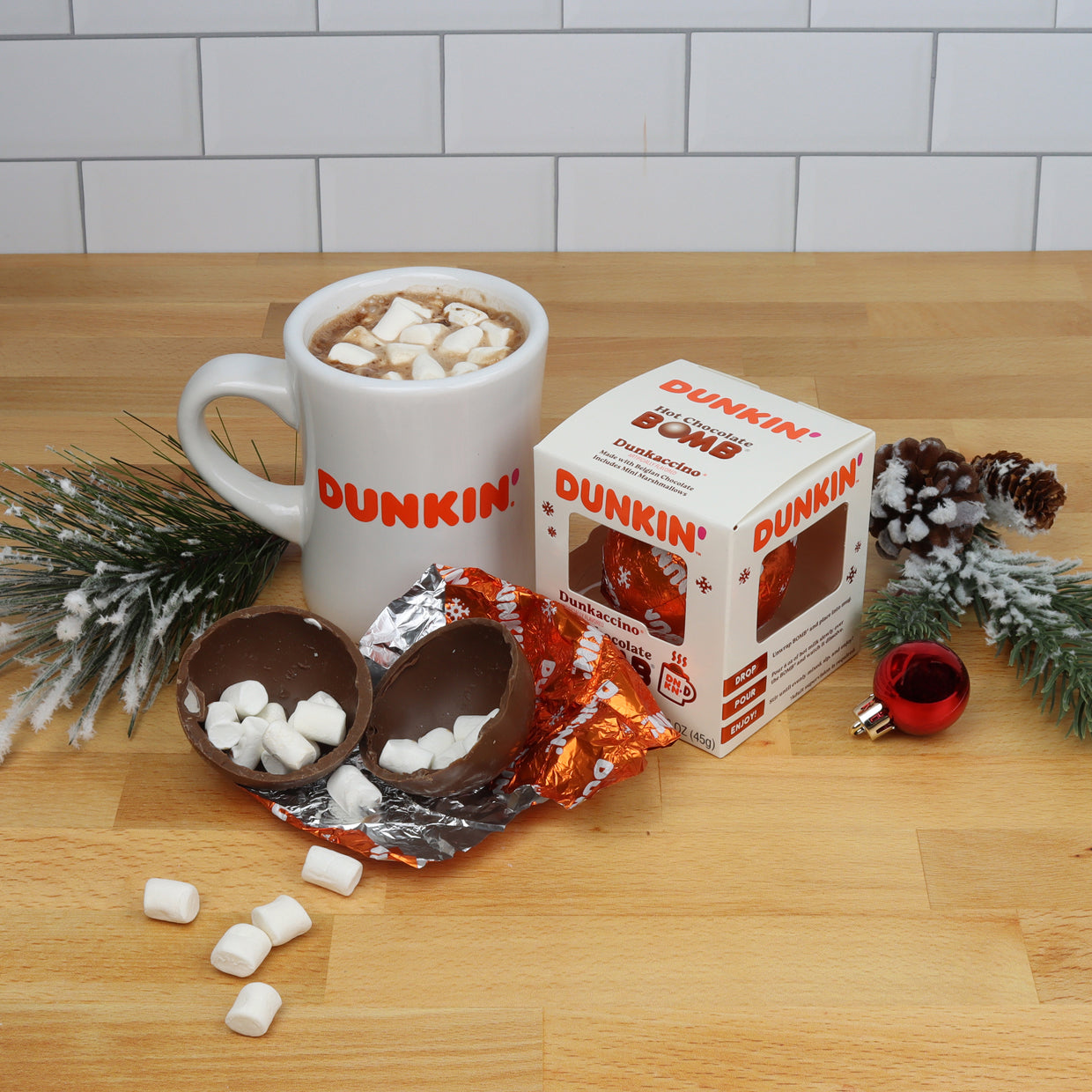 White Dunkin' mug with hot chocolate and mini marshmallows, white Christmas themed box with orange foil with Dunkin' logo wrapped hot chocolate bomb, and open hot chocolate bomb with mini marshmallows