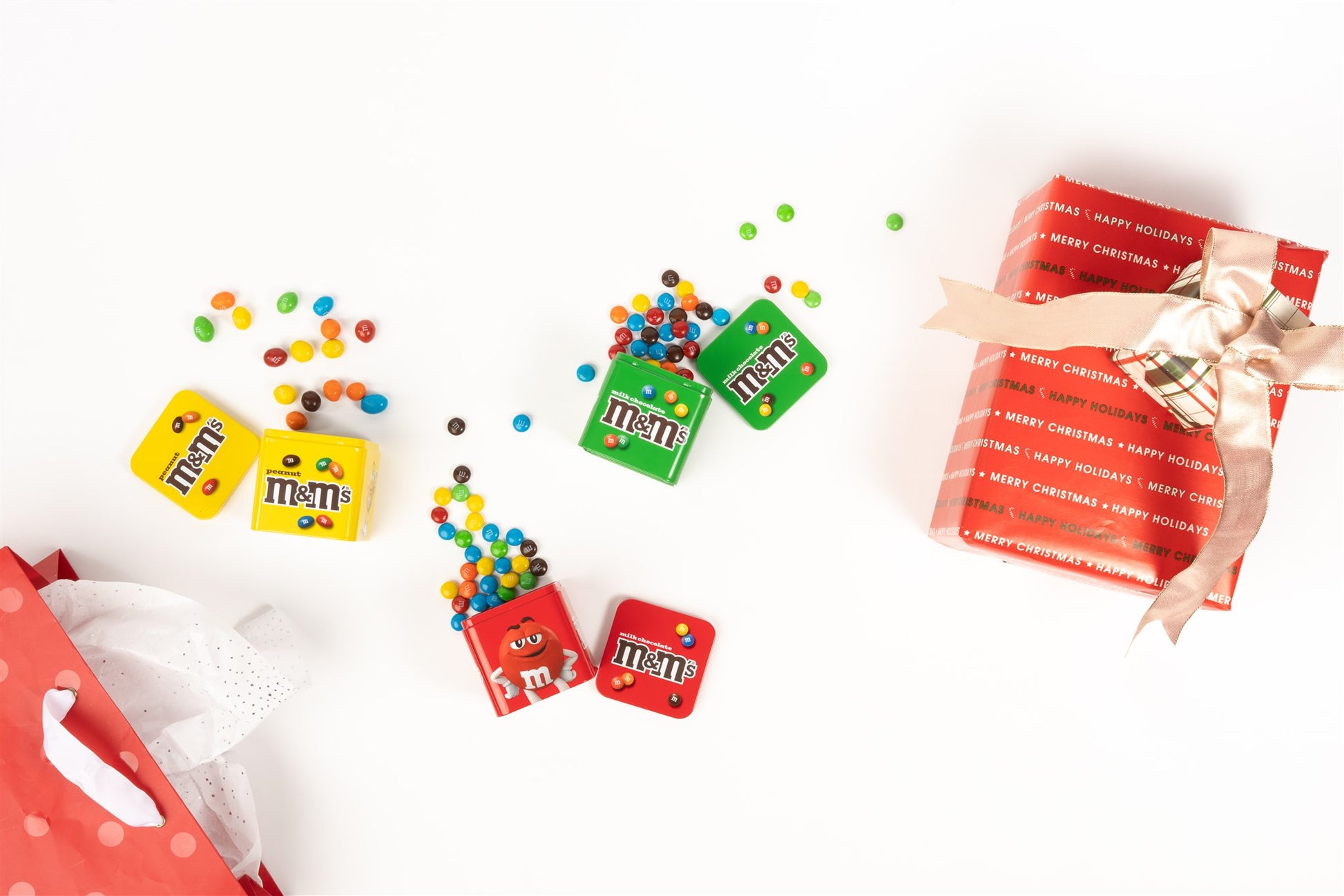 Tinss opened with unwrapped m&m's spilling out of them with a gift bag and present on either side of the tins 