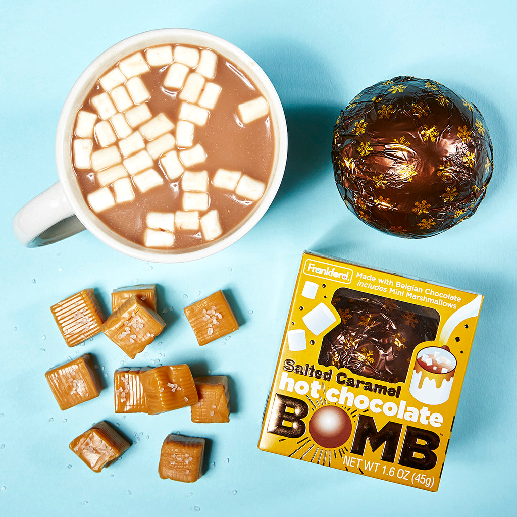 mug with hot chocolate and mini marshmallows, brown foil wrapped hot chocolate bomb, gold box, and salted caramel pieces