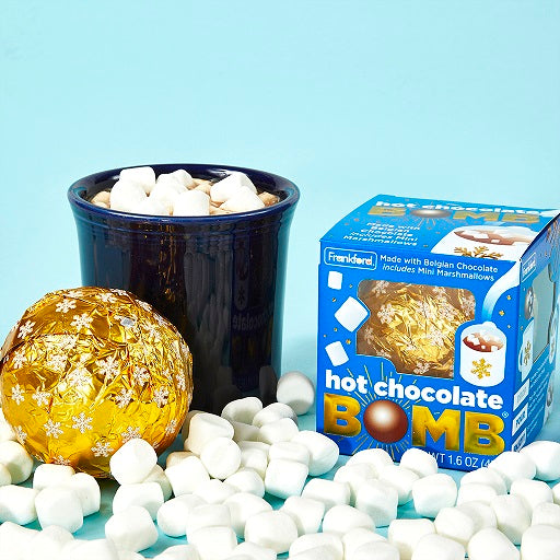 blue hot chocolate bomb box, cup of hot chocolate with mini marshmallows, and a hot chocolate bomb wrapped in gold and snowflake print foil, all surrounded by mini marshmallows