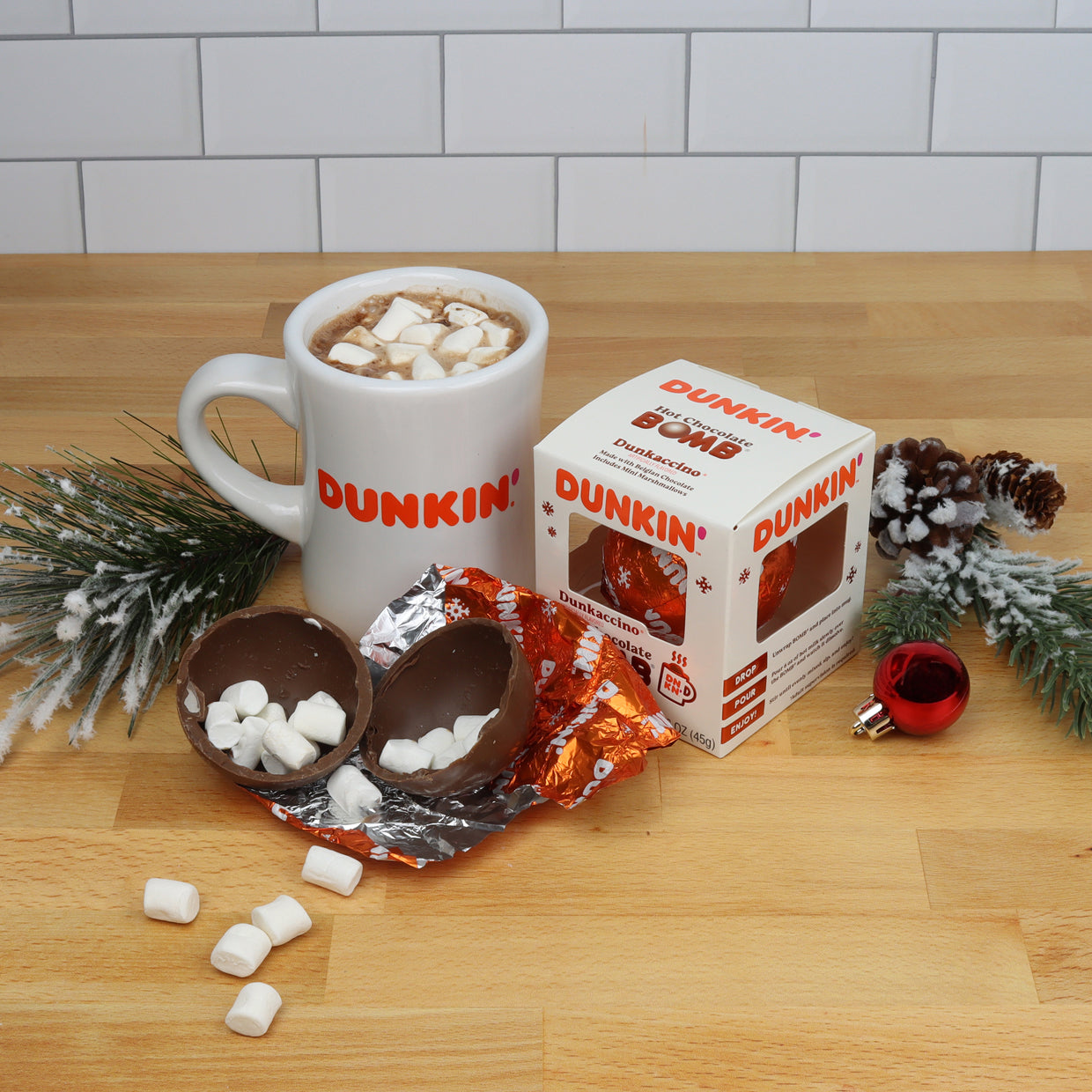 White Dunkin' mug with hot chocolate and mini marshmallows, white Christmas themed box with orange foil with Dunkin' logo wrapped hot chocolate bomb, and open hot chocolate bomb with mini marshmallows 