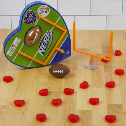 Heart shaped box on its side with toy orange goal post and toy brown football surrounded by red heart shaped gummies