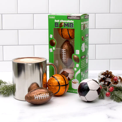 green box with three hot chocolate bombs wrapped and shaped like sports balls with silver mug of hot chocolate